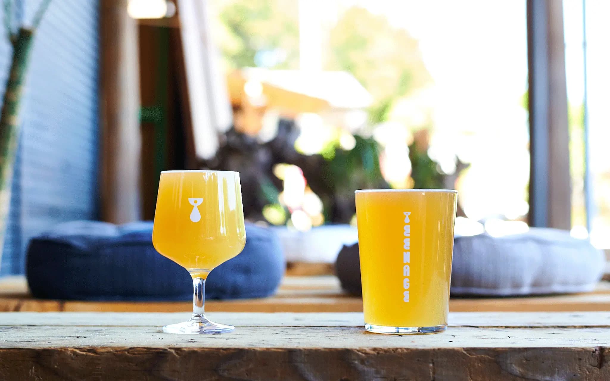 木のテーブルに置かれた2種類のビールグラス、Teenage Brewingのロゴ入りグラスに注がれた濁りのある黄金色のクラフトビール、背景にはテラス席の風景